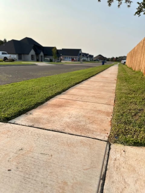 Concrete sidewalk lined with grass and wooden fence running through residential neighborhood with houses and blue sky