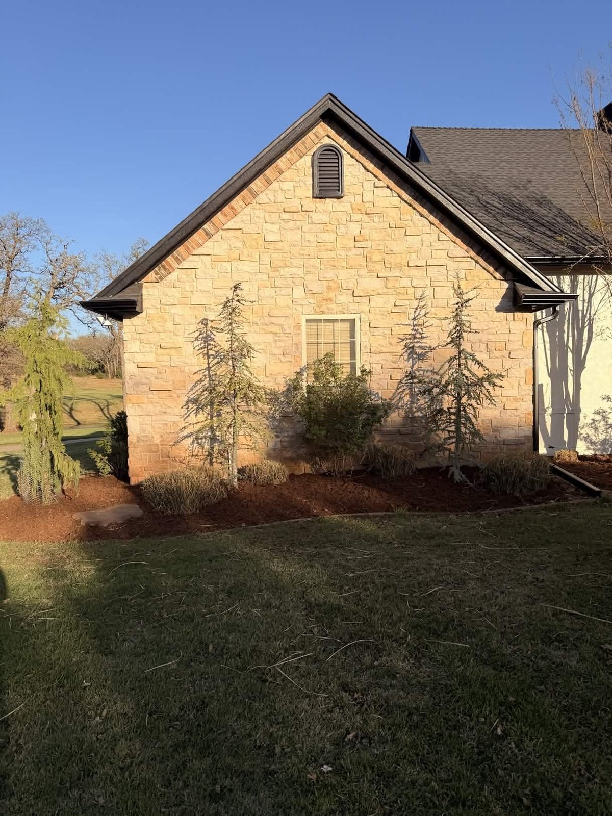Stone house with gabled roof and window, surrounded by dormant shrubs and lawn under clear blue sky