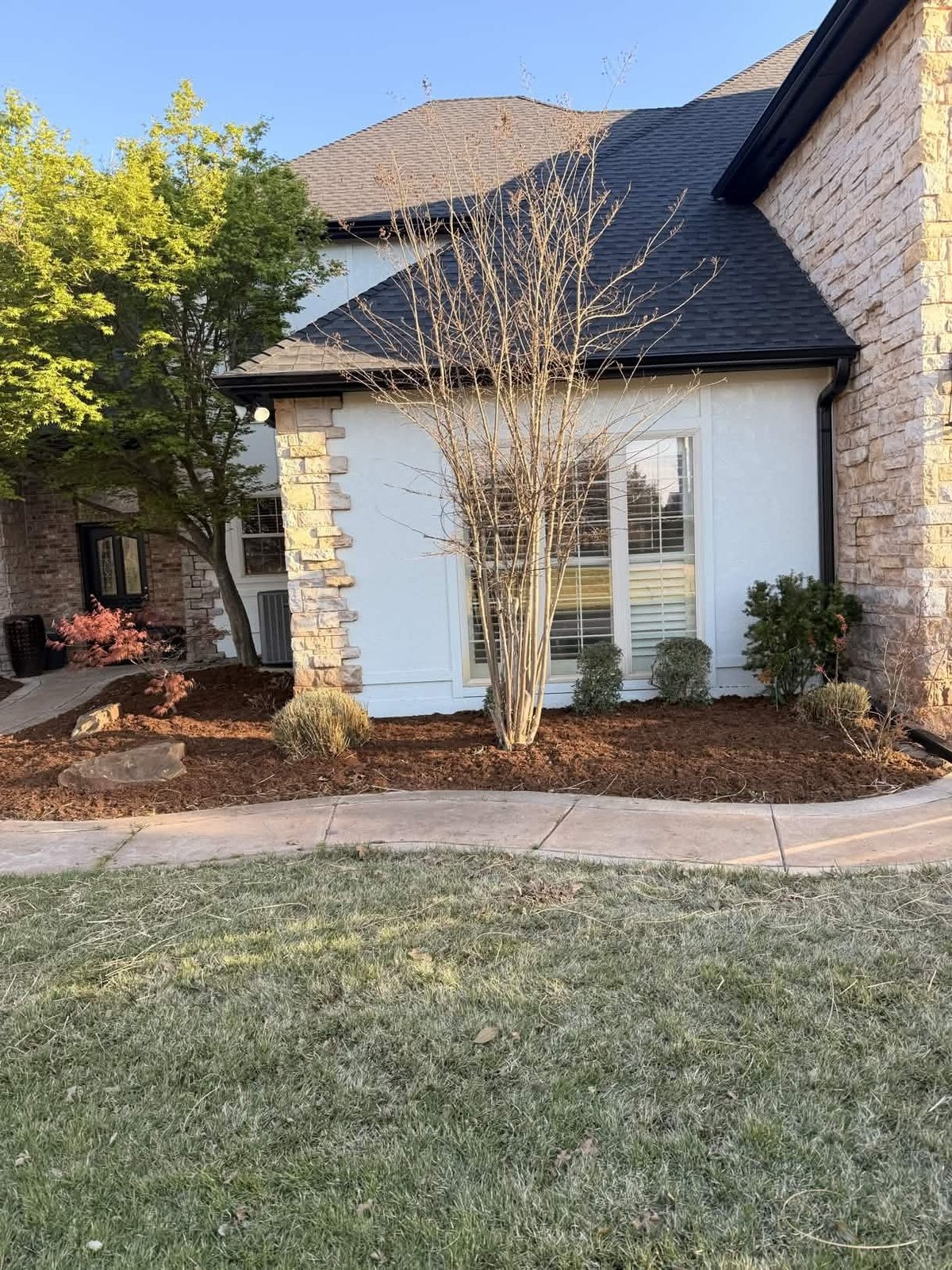Modern brick and white stucco home with dark roof, landscaped yard with bare shrubs, and manicured lawn on a sunny day