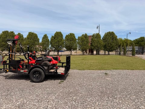 Red and black industrial cart with cargo bed parked on gravel in a park with trees and green lawn in the background